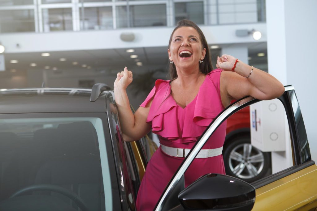 A woman in a pink dress standing next to a car in a showroom, showcasing $1000 Down Payment Cars.