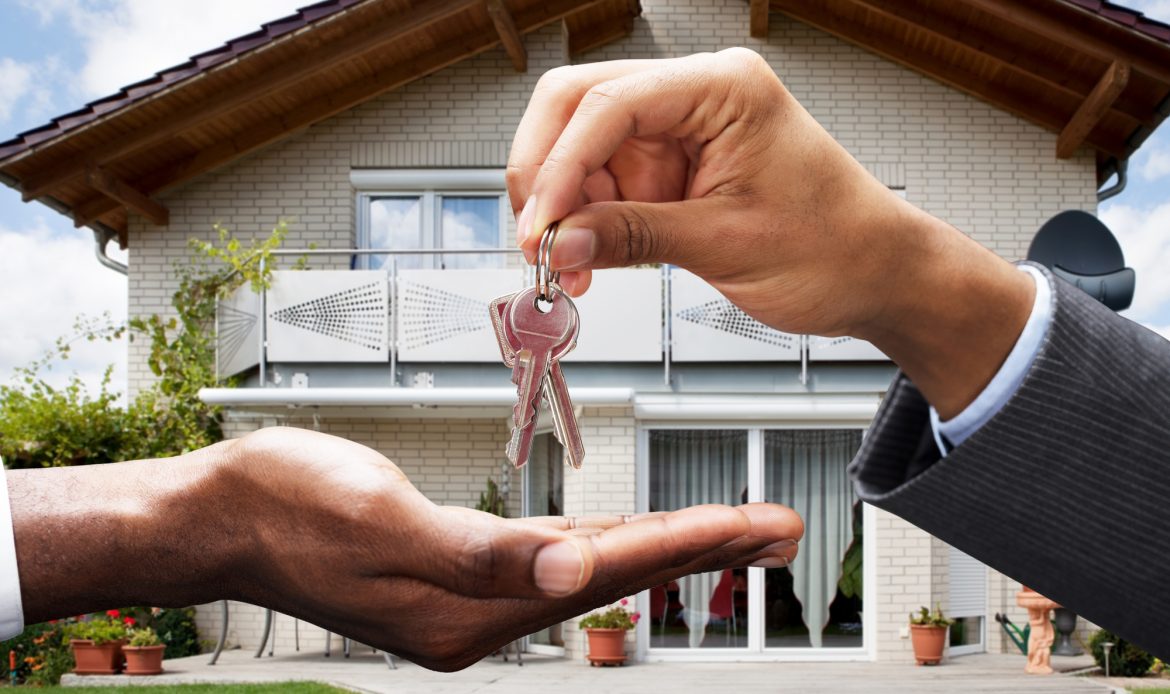 A hand holding a key to a house in front of a house, pondering becoming a real estate agent.
